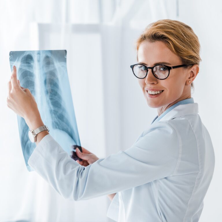 beautiful and cheerful doctor in glasses looking at camera and holding x-ray in clinic